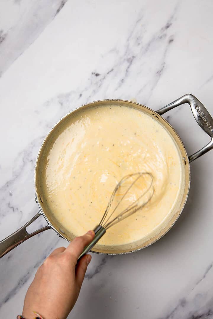 A hand whisking creamy sauce in a stainless steel pan on a marble countertop, preparing the base for a delicious cauliflower and broccoli cheese casserole.