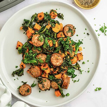 Sliced sheet pan sausage, kale, and roasted sweet potatoes are served on a white plate and garnished with fresh herbs.