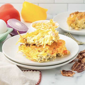 Hashbrown Breakfast Casserole with melted cheese on a white plate, surrounded by eggs, vegetables, and crispy bacon.