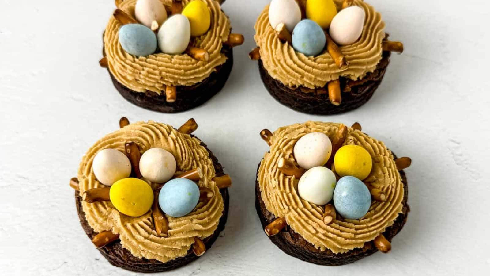 Four chocolate cupcakes topped with brown frosting, candy eggs, and pretzel sticks, arranged on a white surface with scattered colored candy eggs.