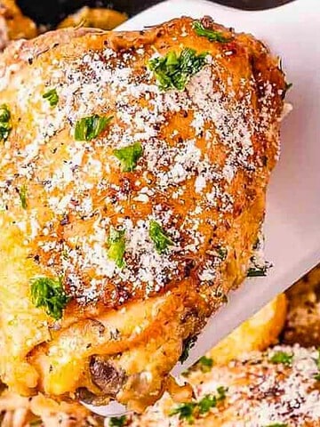 A spatula lifts a seasoned, baked chicken thigh topped with herbs and grated cheese from a pan with more chicken.