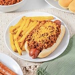 Hot dog topped with savory Hot Dog Chili and shredded cheese, served alongside golden French fries on a white plate—an easy recipe perfect for quick meals.