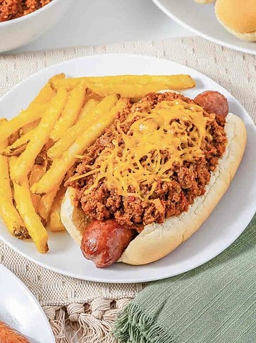 Hot dog topped with savory Hot Dog Chili and shredded cheese, served alongside golden French fries on a white plate—an easy recipe perfect for quick meals.