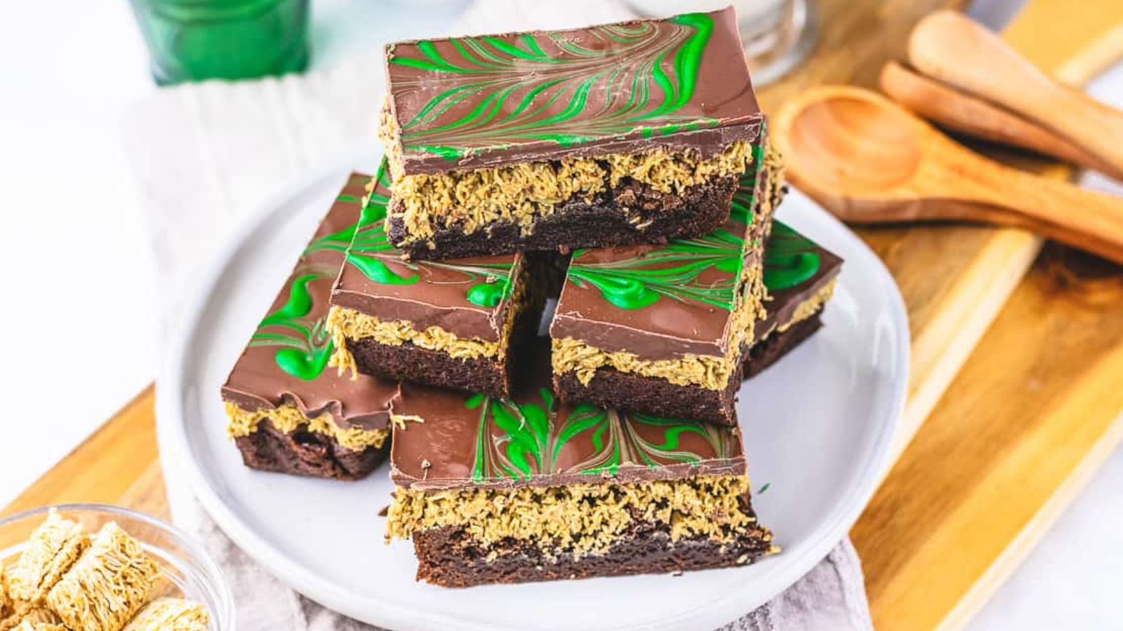 Three chocolate oat bars with green swirled icing are stacked on a white plate, surrounded by baking utensils.