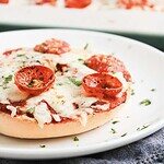 Homemade baked Pepperoni Pizza Bagels on a plate, topped with melted cheese, sliced pepperoni, and sprinkled herbs.