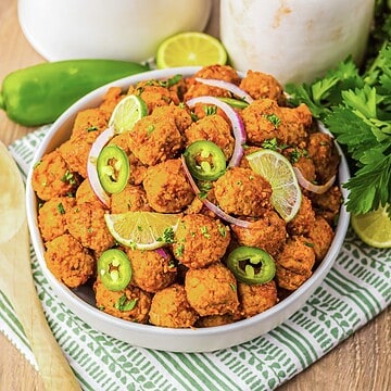 A bowl of spicy meatballs, inspired by birria flavors, garnished with lime slices, jalape&ntilde;os, red onion, and fresh herbs.