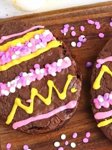 Three egg-shaped brownies decorated with pink and yellow icing and sprinkles on a wooden board.