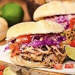 Slow cooker Tex Mex pulled pork sandwich topped with red cabbage slaw and tomatoes on a bun, served with lime wedges on the side.