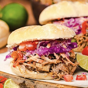 Slow cooker Tex Mex pulled pork sandwich topped with red cabbage slaw and tomatoes on a bun, served with lime wedges on the side.