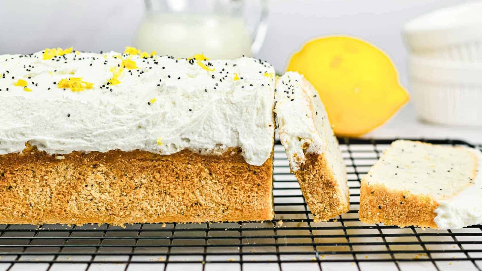 Lemon poppy seed loaf with white frosting, lemon zest, and a sliced lemon on a cooling rack.