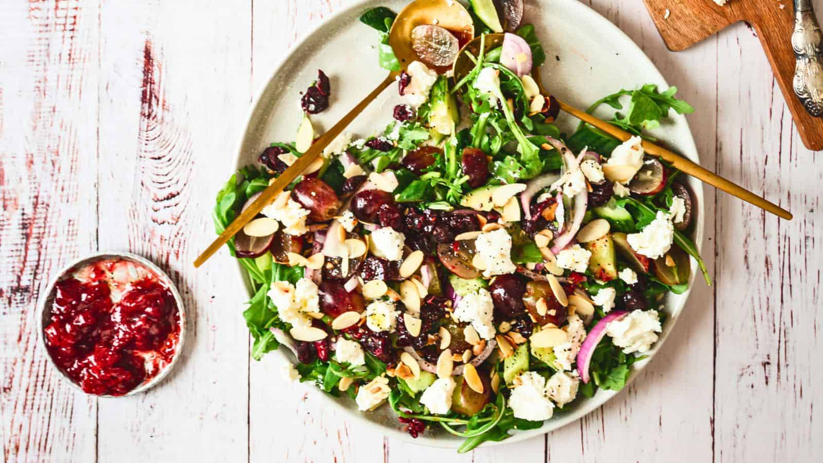 A goat cheese salad is served on a round white plate. The salad includes mixed greens, sliced grapes, crumbled goat cheese, sliced red onions, and almond slivers. A gray cloth is placed beside the plate on a white wooden surface. There is a wooden cutting board with a knife in the background.