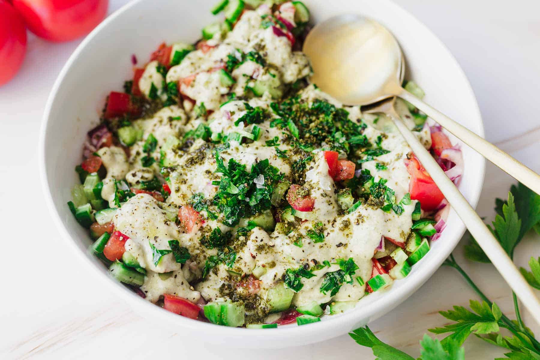 A white bowl filled with an Israeli salad composed of chopped cucumbers, tomatoes, onions, and herbs, topped with a creamy dressing and sprinkled with spices. Two gold-colored spoons rest in the bowl. Fresh parsley and two whole tomatoes are placed beside this one serving.
