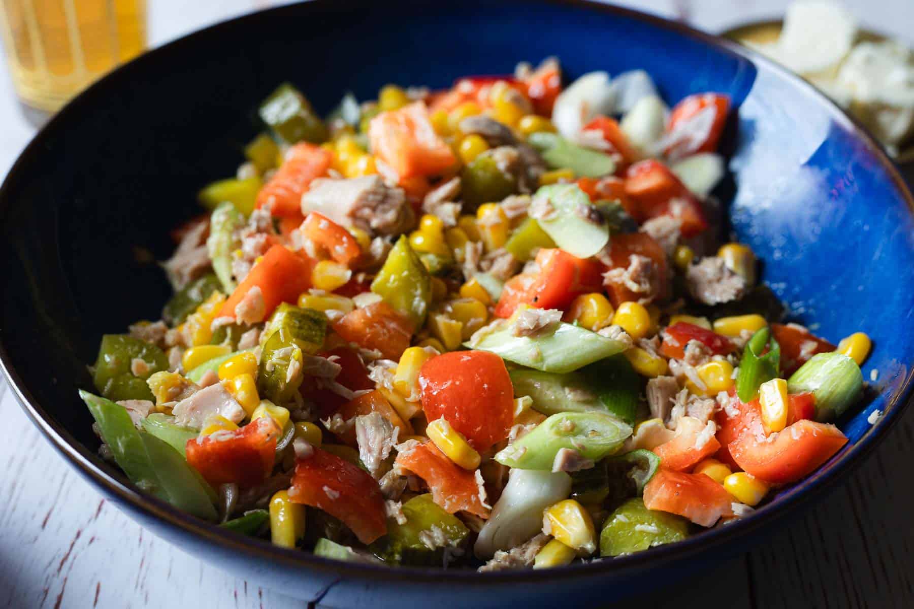 A blue bowl filled with a colorful salad of tomatoes, corn, green onions, tuna, and pickles.