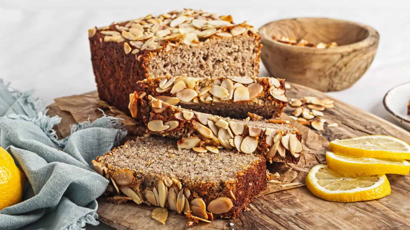 Sliced almond bread on a wooden board with lemon slices and a bowl of sliced almonds in the background.