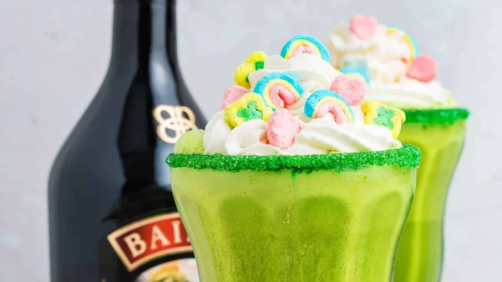 A tall glass of milkshake topped with whipped cream and colorful cereal marshmallows sits on a surface with additional marshmallows scattered around it.