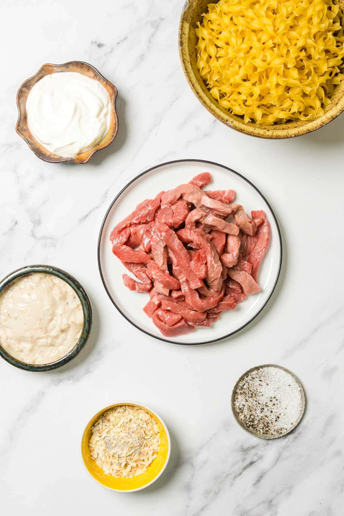 Ingredients for beef stroganoff arranged on a white marble surface, including beef strips, noodles, and seasonings.