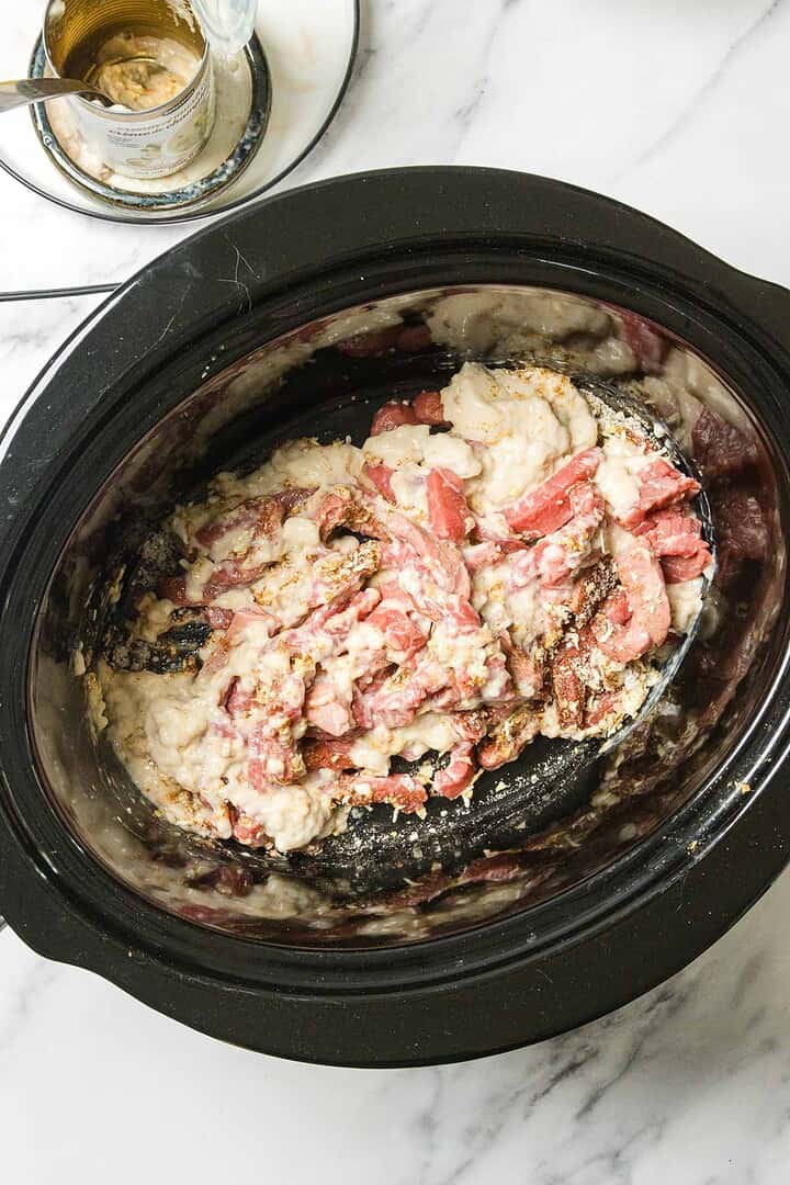 Raw beef strips mixed with a creamy sauce in a black slow cooker on a marble countertop.