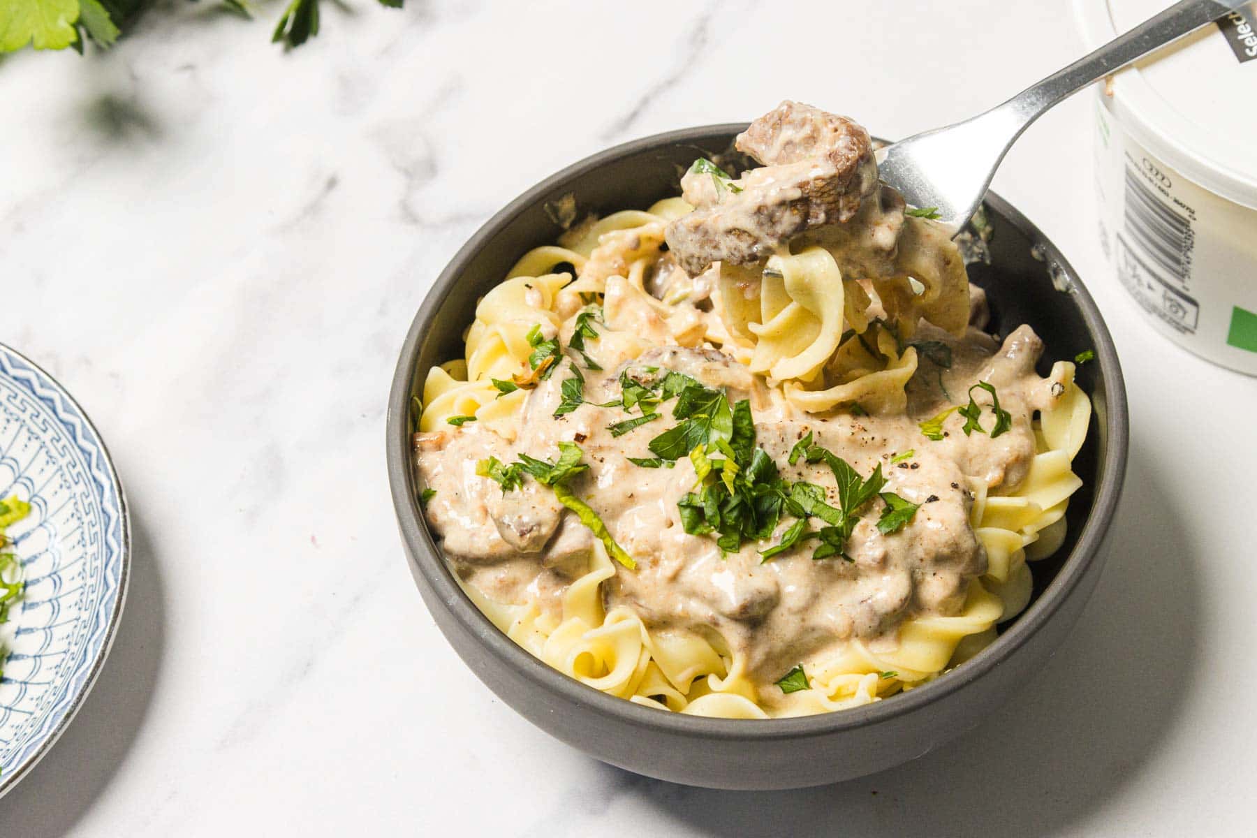 Bowl of creamy beef stroganoff over egg noodles, garnished with chopped herbs, on a marble surface.