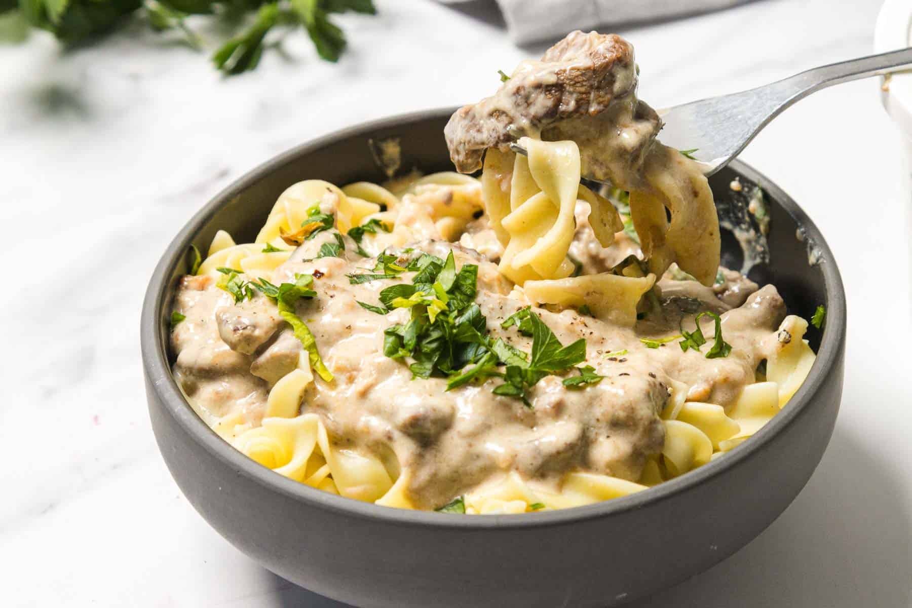 A bowl of creamy beef stroganoff over egg noodles, garnished with chopped parsley, with a forkful lifted.
