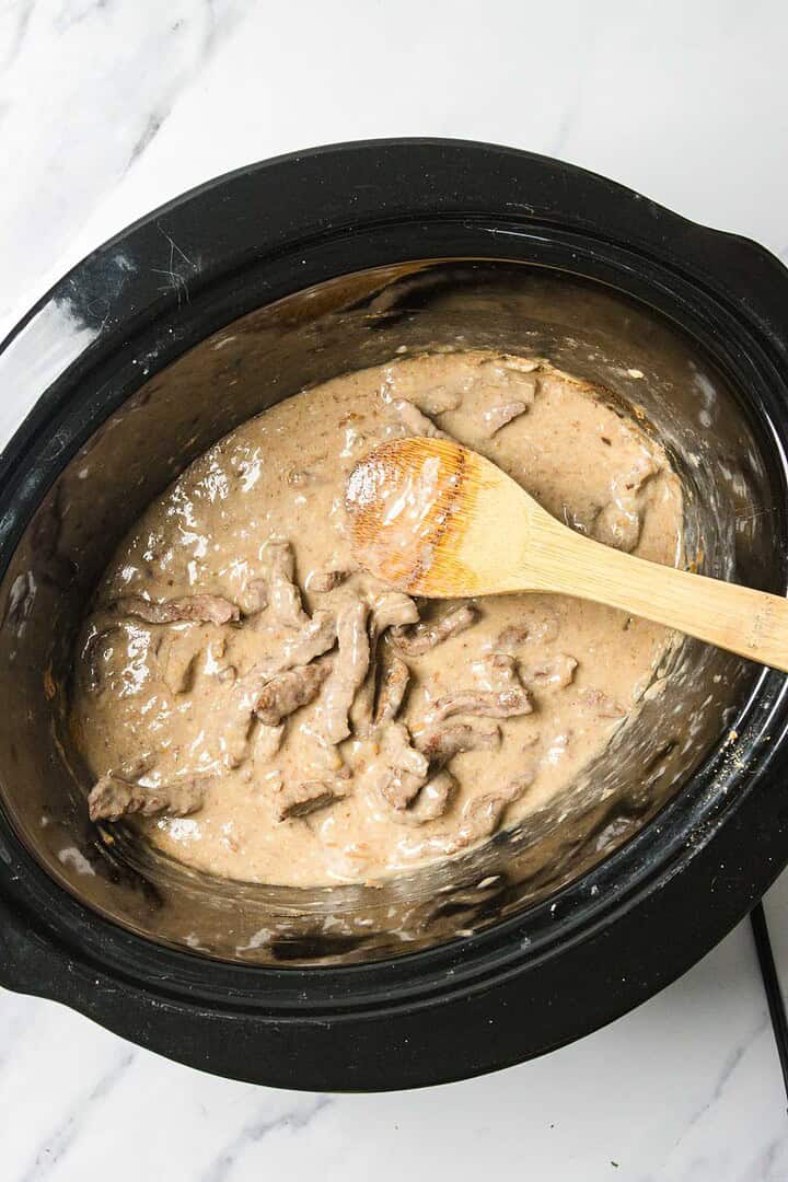 Beef stroganoff in a slow cooker with a wooden spoon resting on top of the creamy mixture.