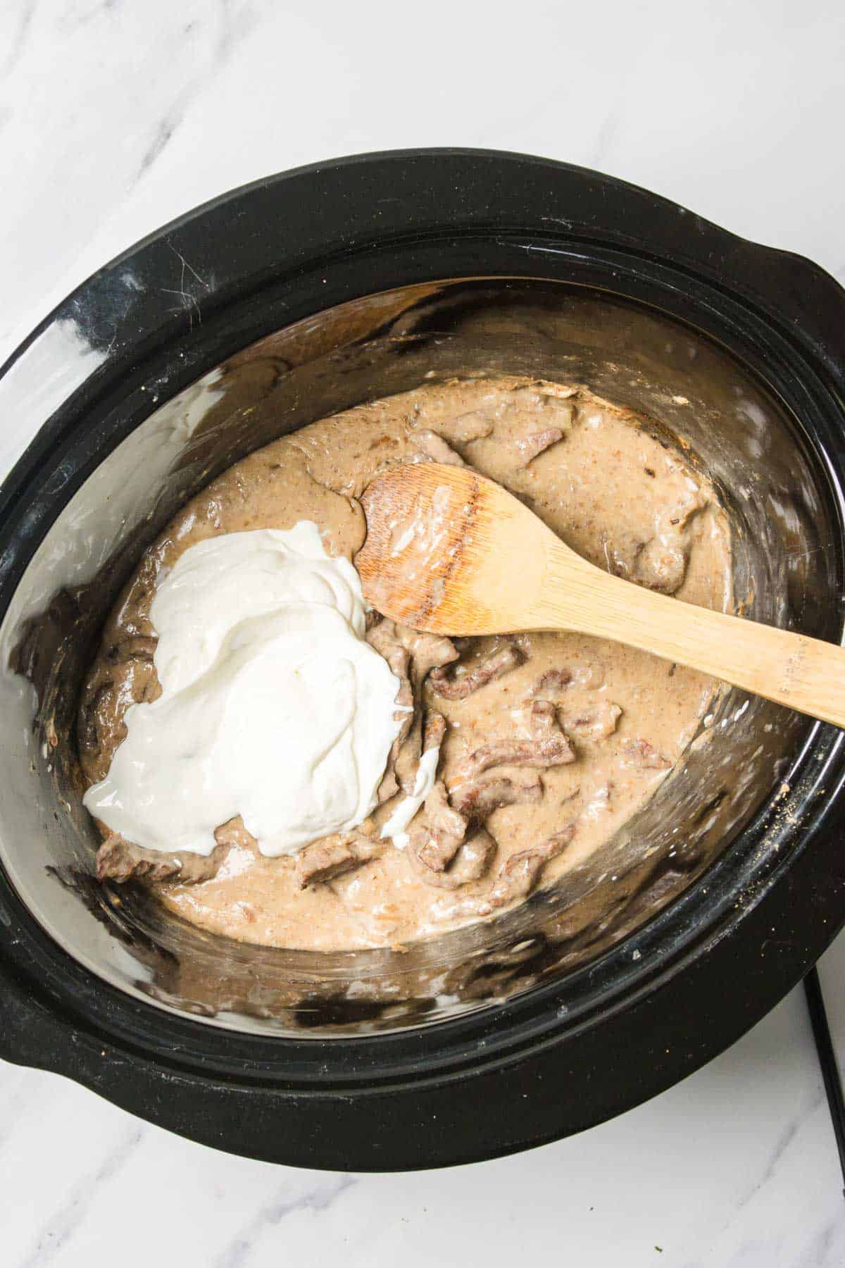 Beef stroganoff in a slow cooker with a dollop of sour cream and a wooden spoon on top.