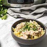 A bowl of creamy slow cooker beef stroganoff with parsley sits in front of the appliance on a marble surface.