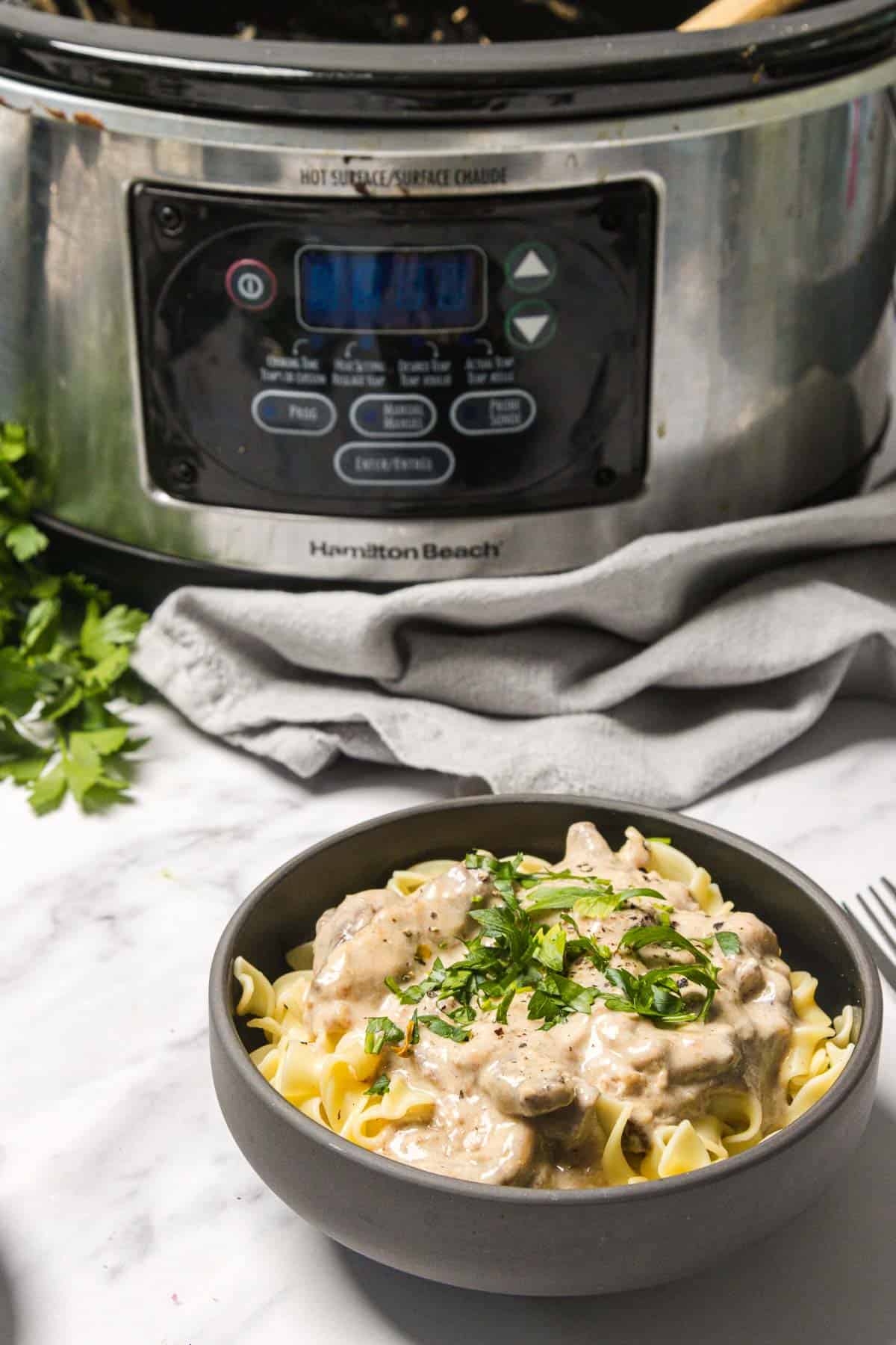 A bowl of creamy slow cooker beef stroganoff with parsley sits in front of the appliance on a marble surface.