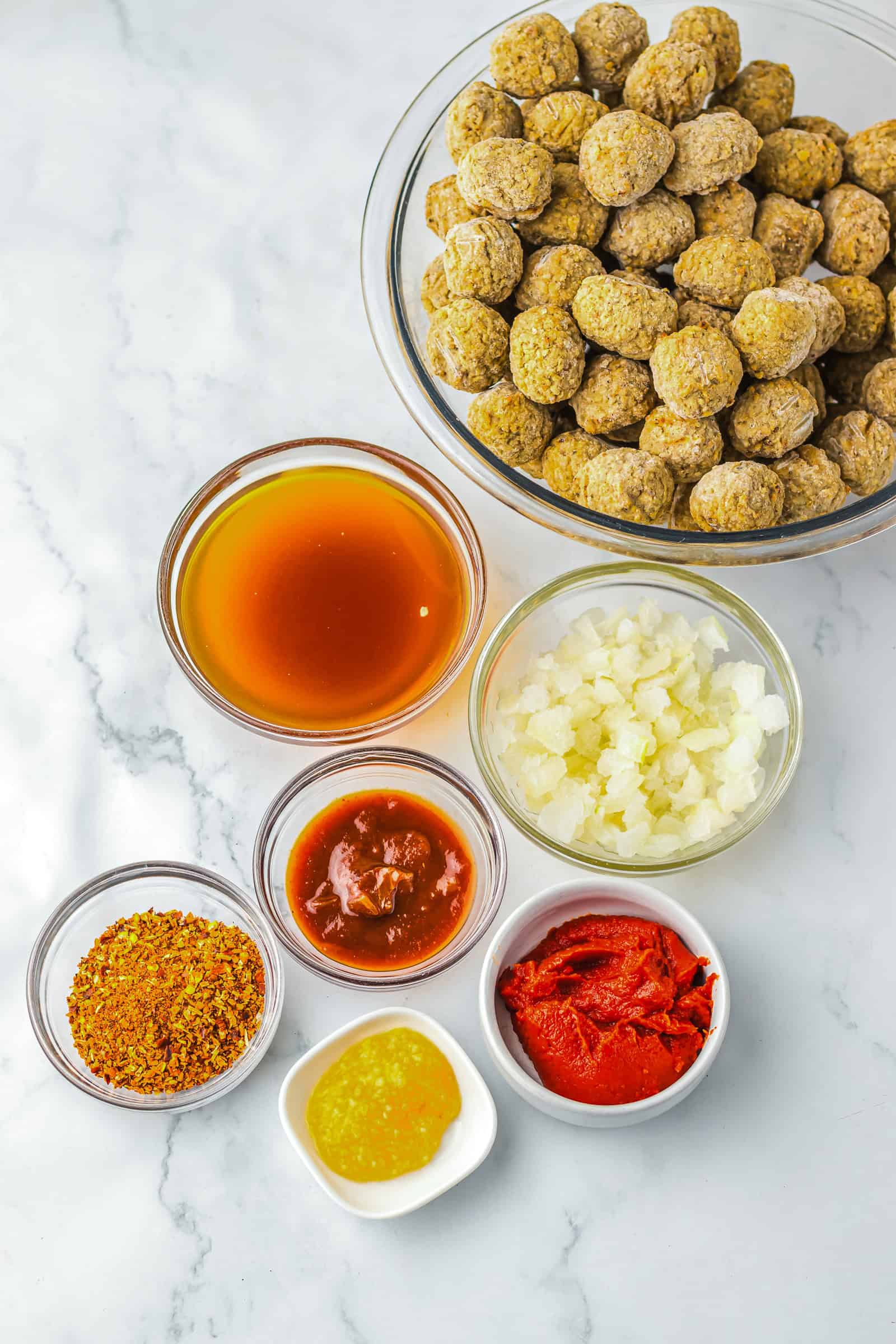 Bowls of Birria Meatballs, chopped onions, tomato paste, sauce, broth, yellow sauce, and seasoning on a marble surface. Perfect for making flavorful meatballs in your slow cooker.