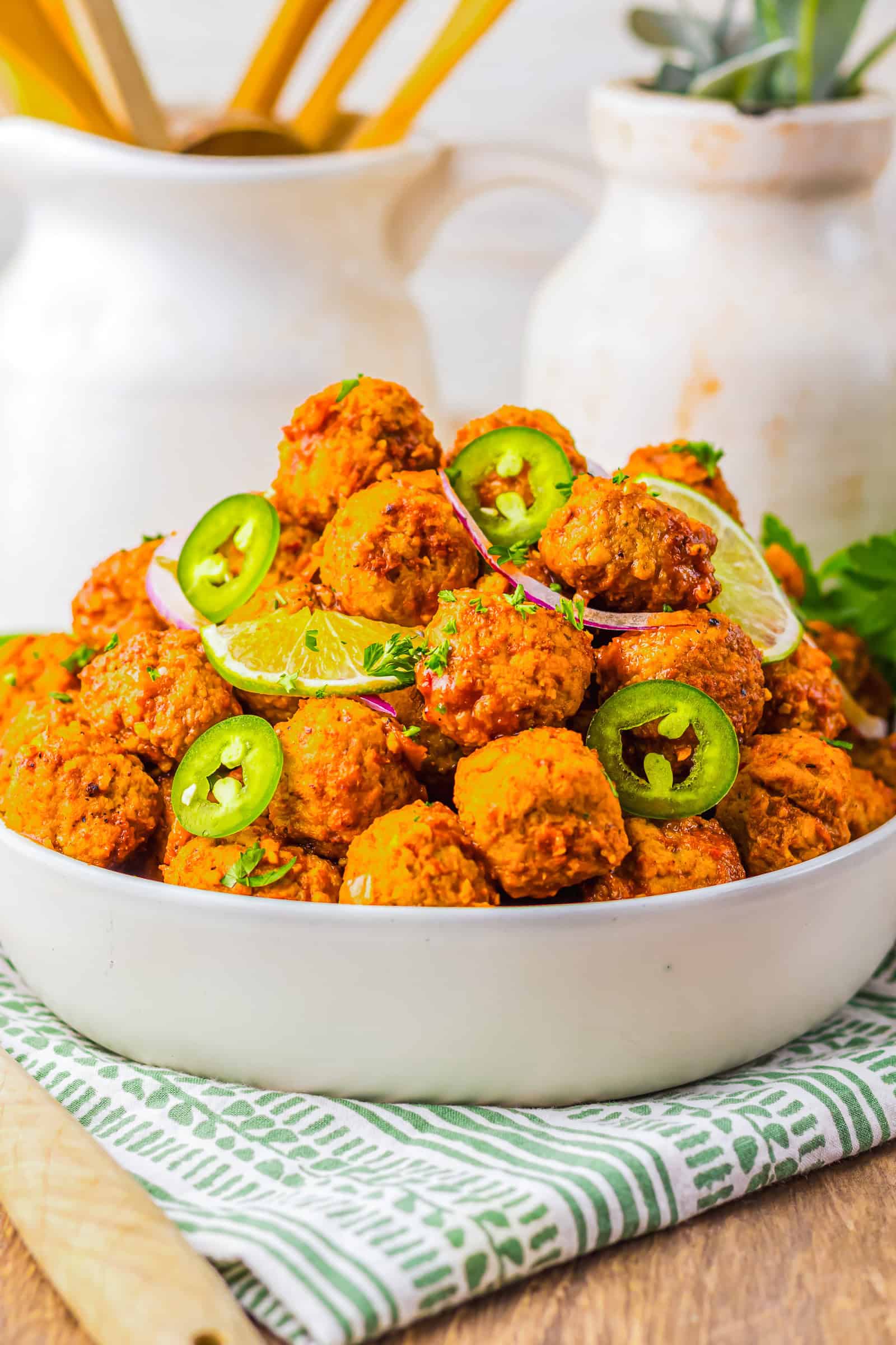 A bowl of spicy birria meatballs garnished with lime slices, jalape&ntilde;os, and herbs rests on a patterned napkin. Perfect for preparing in a slow cooker for rich, bold flavor.
