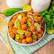 A bowl of spicy falafel garnished with lime, jalapeño slices, and red onion sits alongside herbs and utensils—perfect inspiration for making Birria Meatballs in your slow cooker.