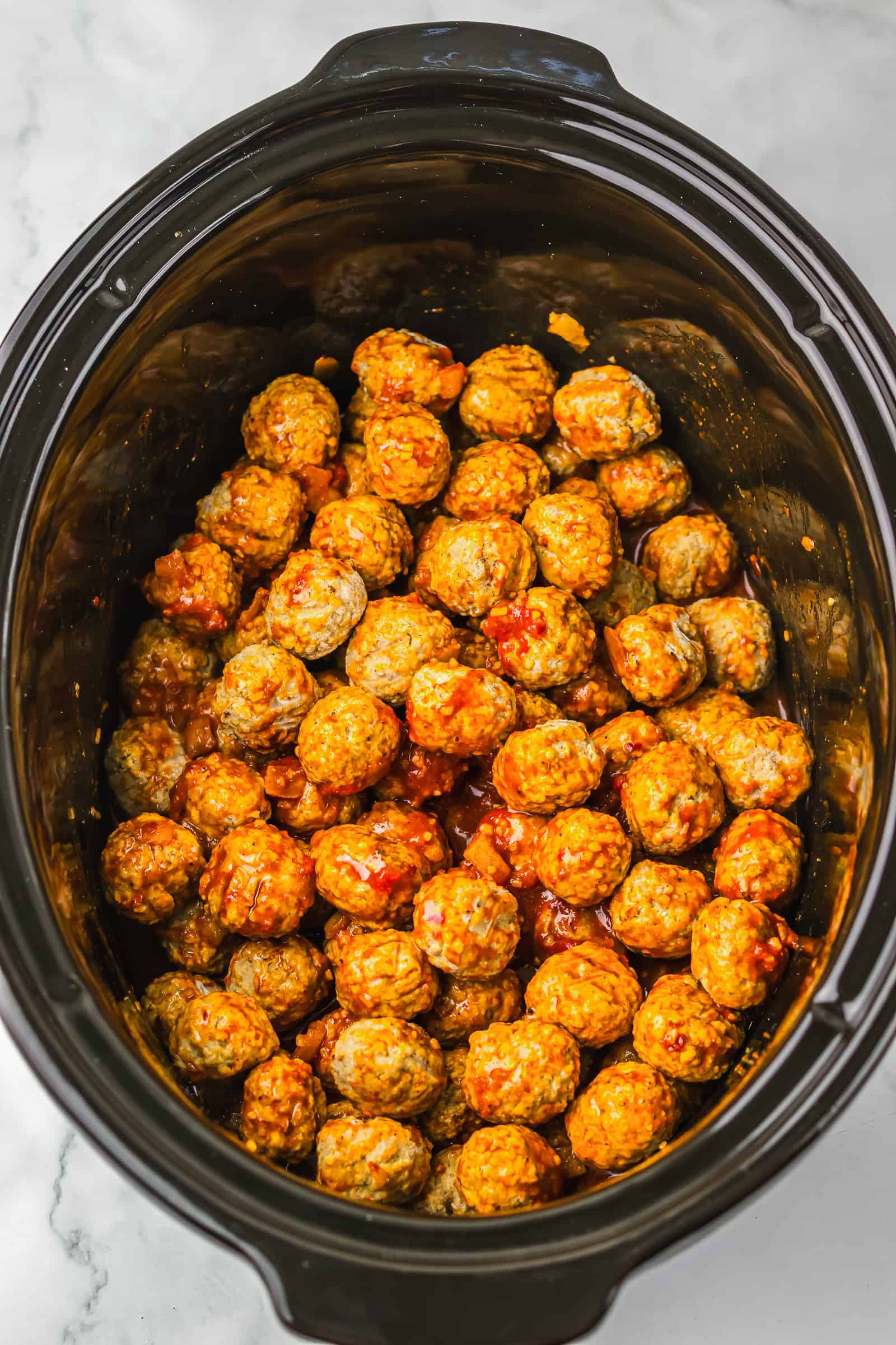 Slow cooker filled with saucy birria meatballs on a white marble countertop.