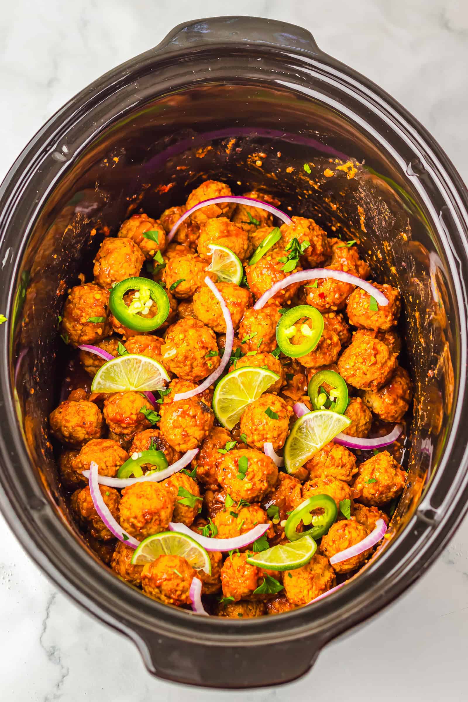 Birria-inspired meatballs in a slow cooker, topped with jalape&ntilde;o slices, lime wedges, and red onion.