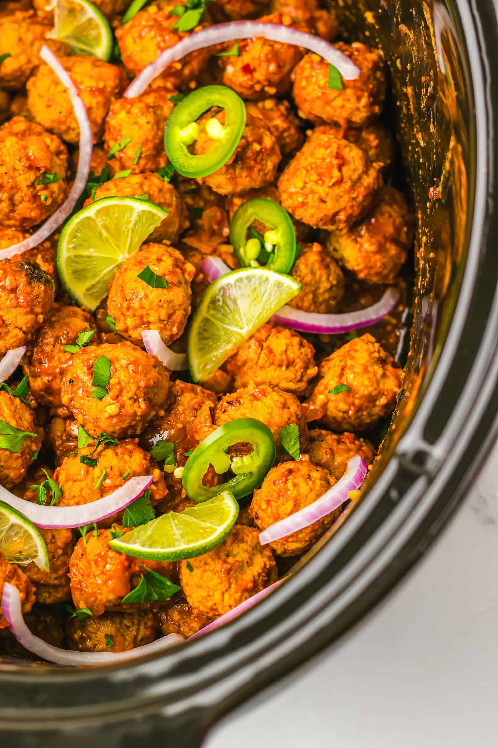 Birria-inspired spicy meatballs simmer in a slow cooker, topped with lime slices, jalape&ntilde;o, red onion, and fresh herbs.