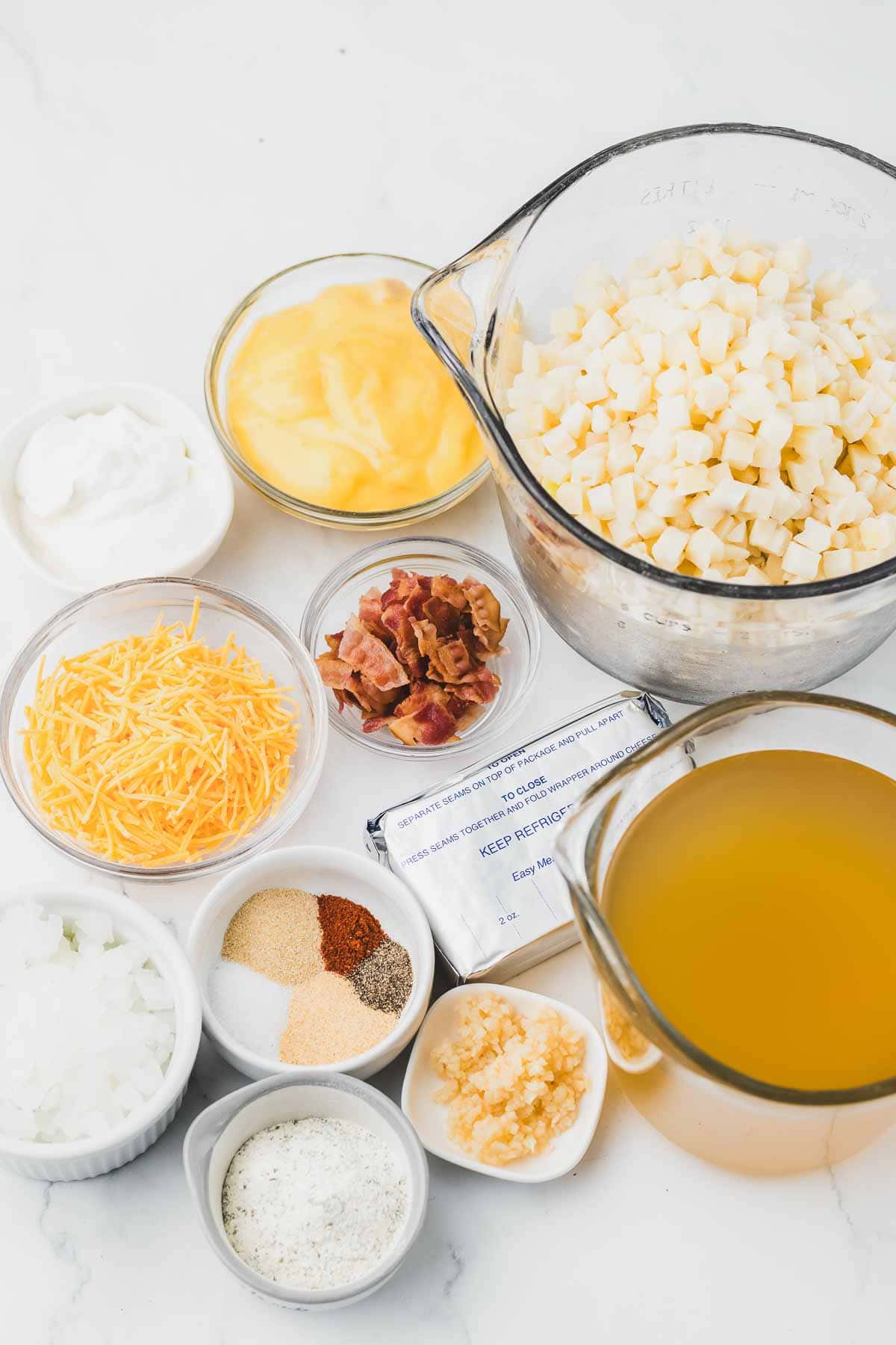 Various bowls with diced potatoes, broth, cheese, bacon, sour cream, and seasonings on a white surface—perfect ingredients for a comforting Crack Potato Soup or classic Slow Cooker Potato Soup.