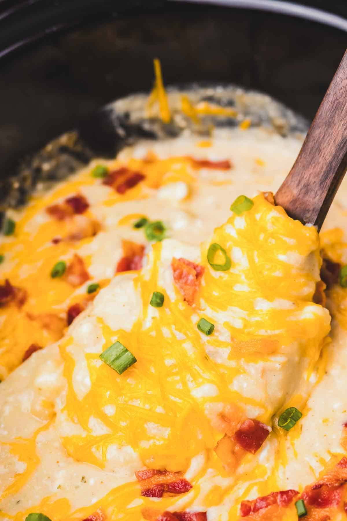 Spoon scooping cheesy, creamy crack potato soup topped with bacon and green onions from a slow cooker.