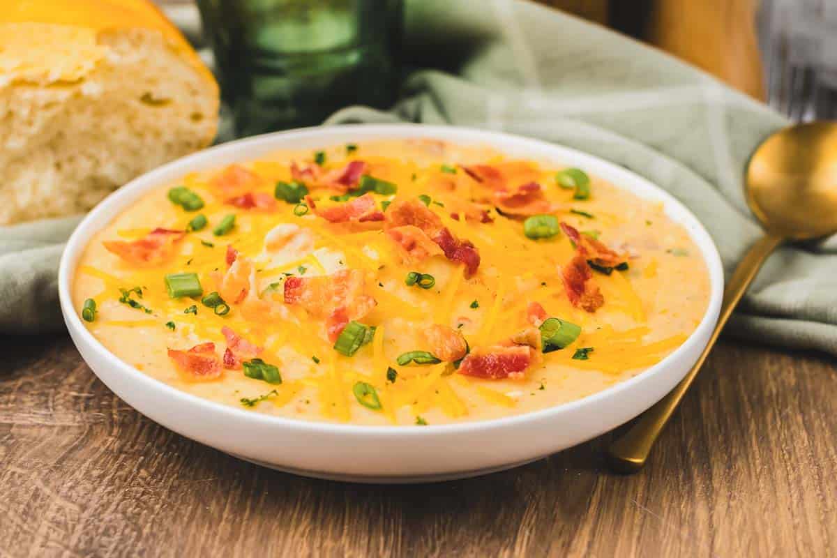 Bowl of creamy slow cooker potato soup topped with shredded cheese, crispy bacon pieces, and chopped green onions on a table.