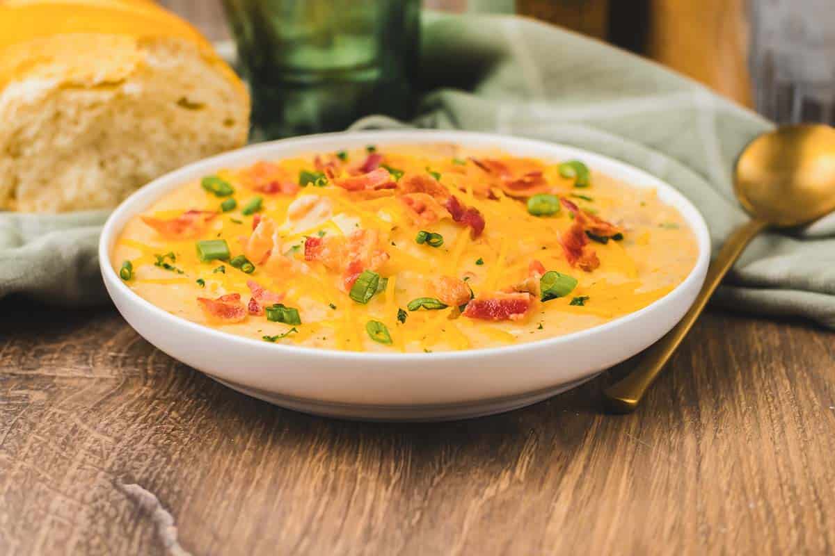 Bowl of slow cooker potato soup loaded with cheese, bacon, and green onions, with fresh bread in the background.