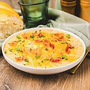 Bowl of creamy slow cooker crack potato soup topped with bacon, green onions, and cheese, served with bread and glasses in the background.