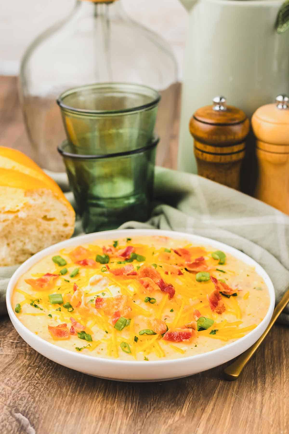 A bowl of cheesy Crack Potato Soup topped with bacon and green onions, served with bread, glasses, and salt and pepper shakers nearby.