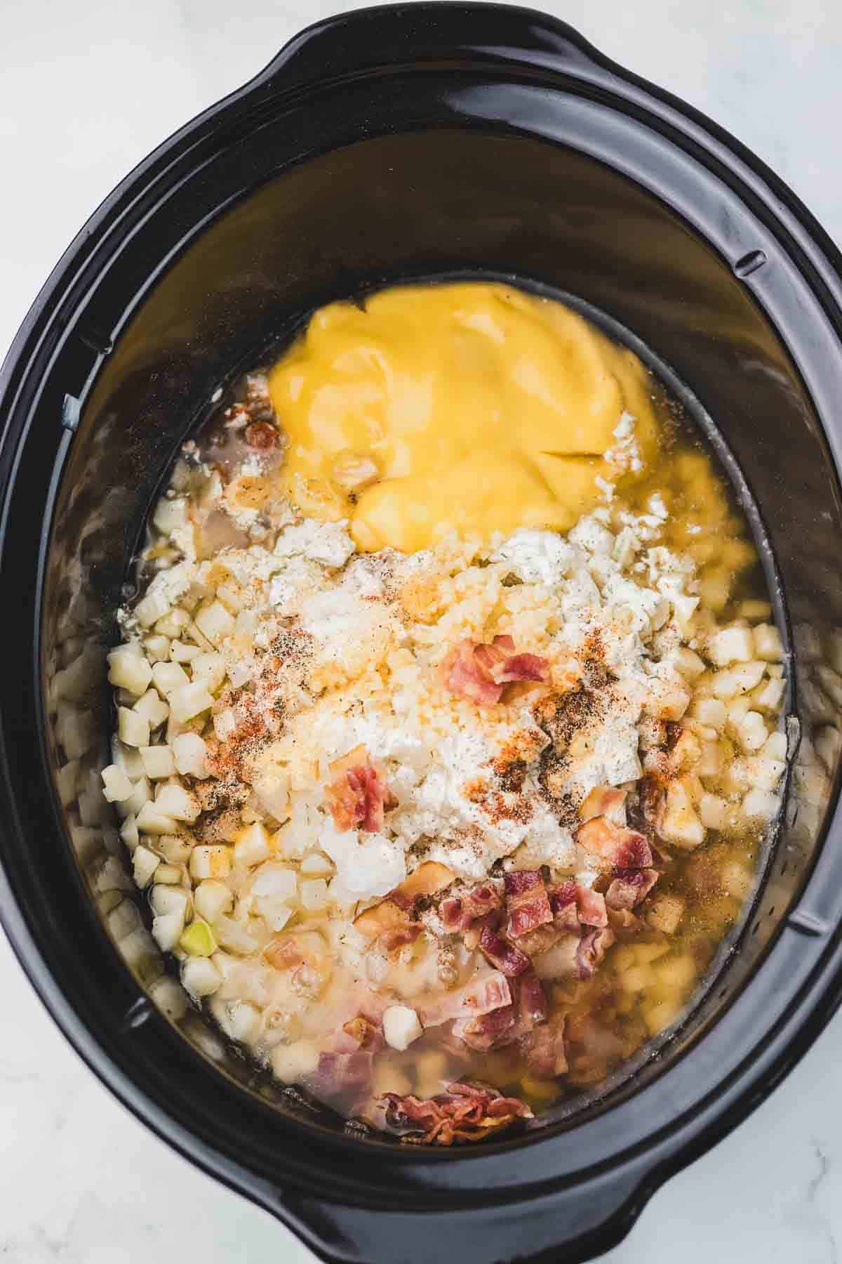 Crockpot filled with diced potatoes, bacon, shredded cheese, seasoning, and soup ready to be mixed for a tasty Slow Cooker Crack Potato Soup.