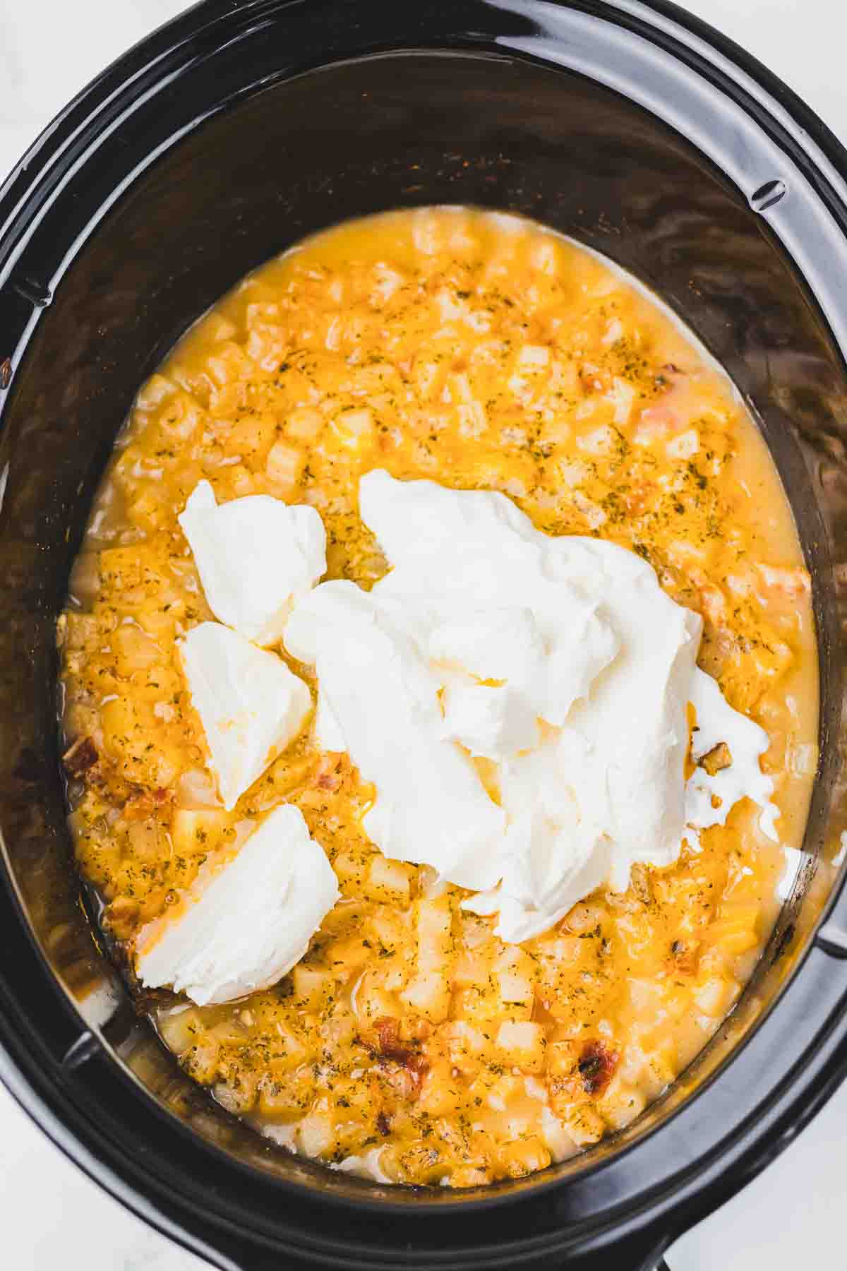 Slow Cooker filled with cheesy Crack Potato Soup, topped with dollops of cream cheese before mixing.