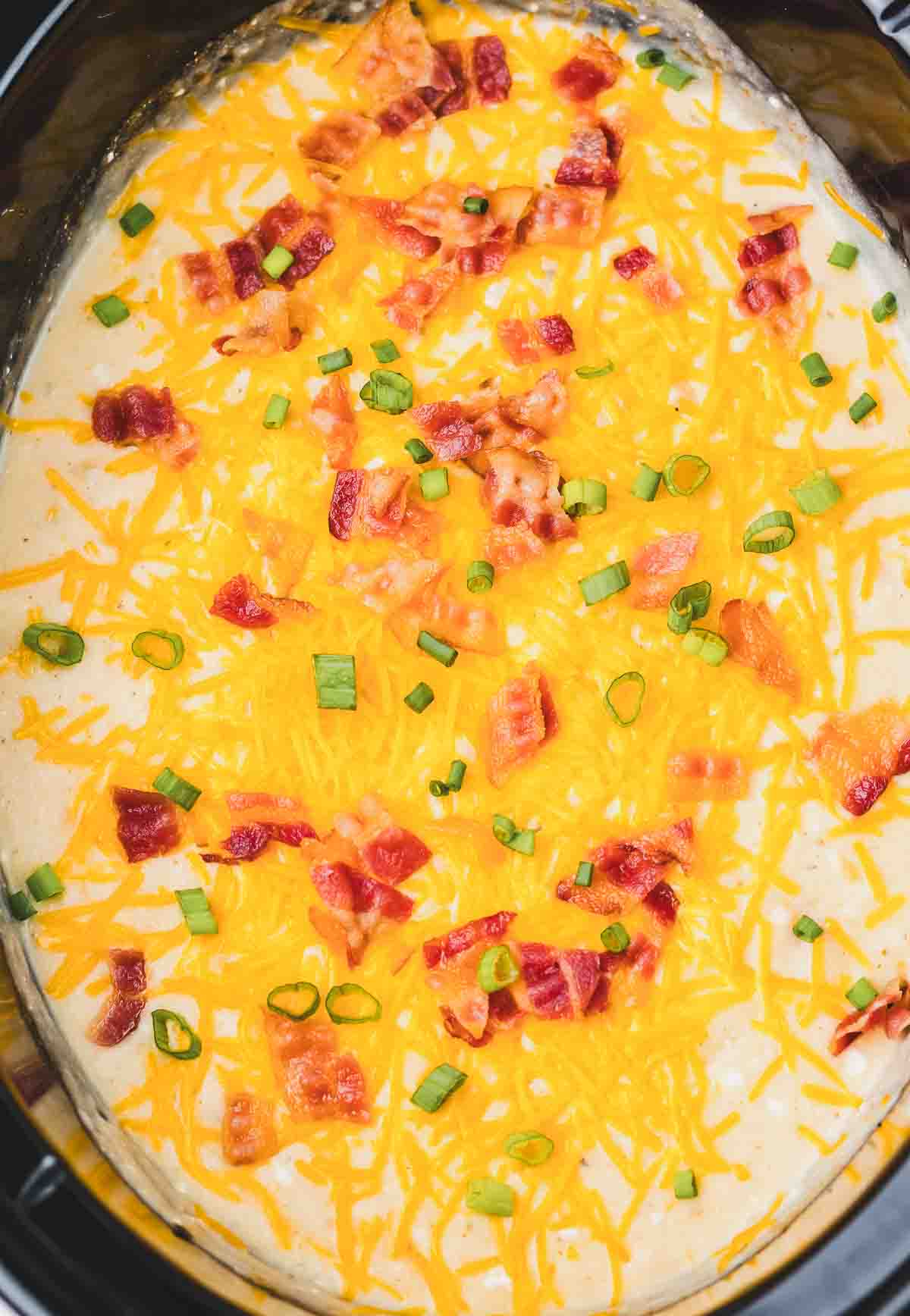 Creamy Crack Potato Soup in a slow cooker, topped with shredded cheddar cheese, crispy bacon pieces, and sliced green onions.