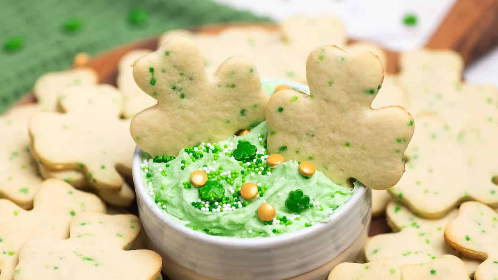 Shamrock-shaped cookies with green frosting and sprinkles in a bowl, surrounded by more cookies.