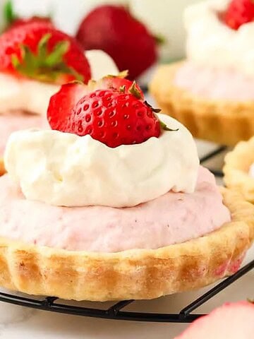 Mini strawberry tarts topped with whipped cream and fresh strawberry halves on a cooling rack.