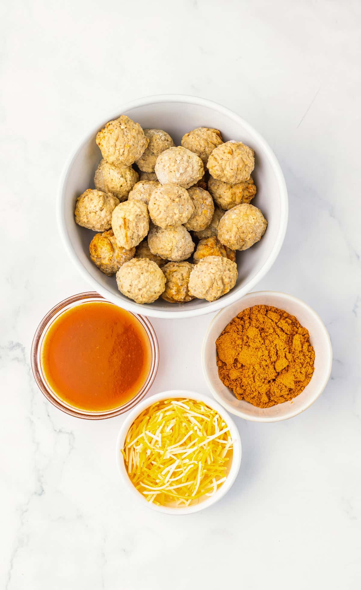 Overhead view of a bowl of 4 ingredient enchilada meatballs, with bowls of sauce, shredded cheese, and a spice mix on a white surface.