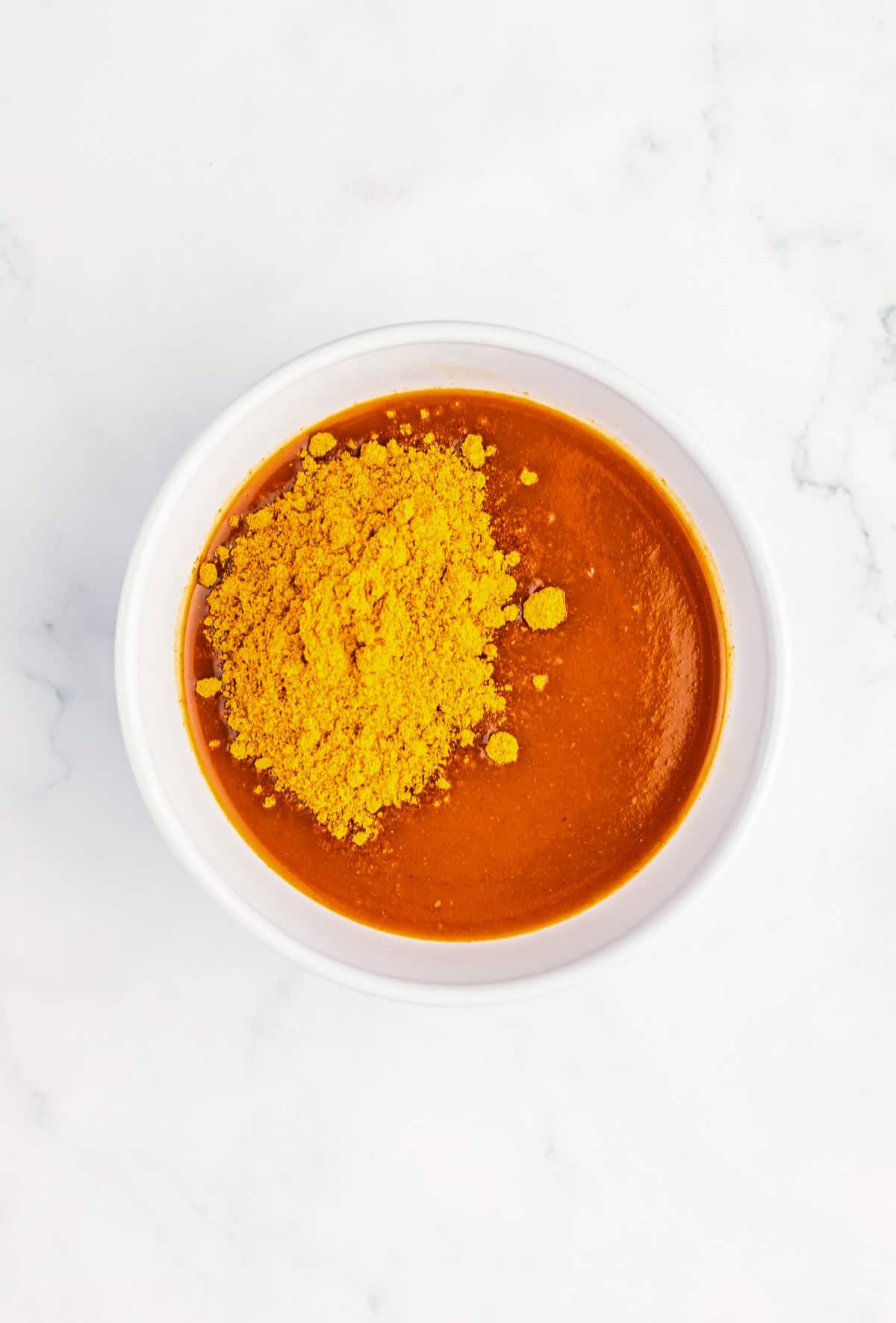 A white bowl with red sauce, topped with a heap of yellow powdered seasoning, holds savory Enchilada Meatball goodness&mdash;an easy 4 ingredient meal on a white marble surface.