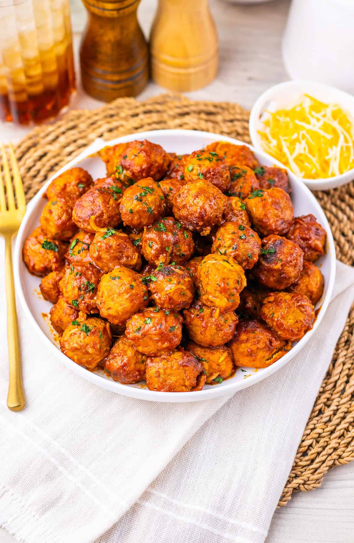 A bowl of saucy Enchilada Meatball garnished with herbs, beside shredded cheese and a fork on a napkin&mdash;an easy 4 Ingredient Recipe perfect for your slow cooker.