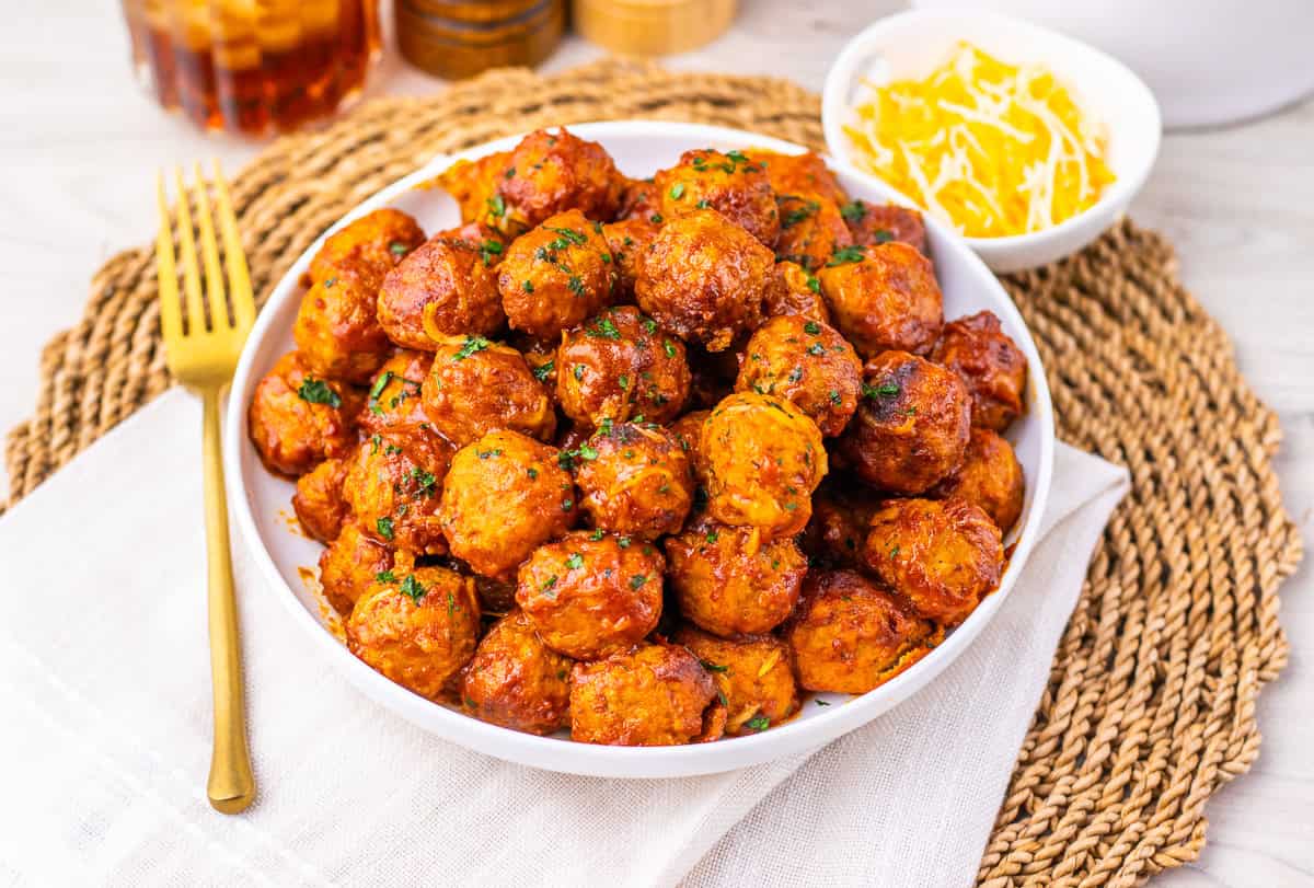 A bowl of 4 ingredient enchilada meatballs glazed to perfection and garnished with herbs, next to a small dish of shredded cheese on a woven placemat.