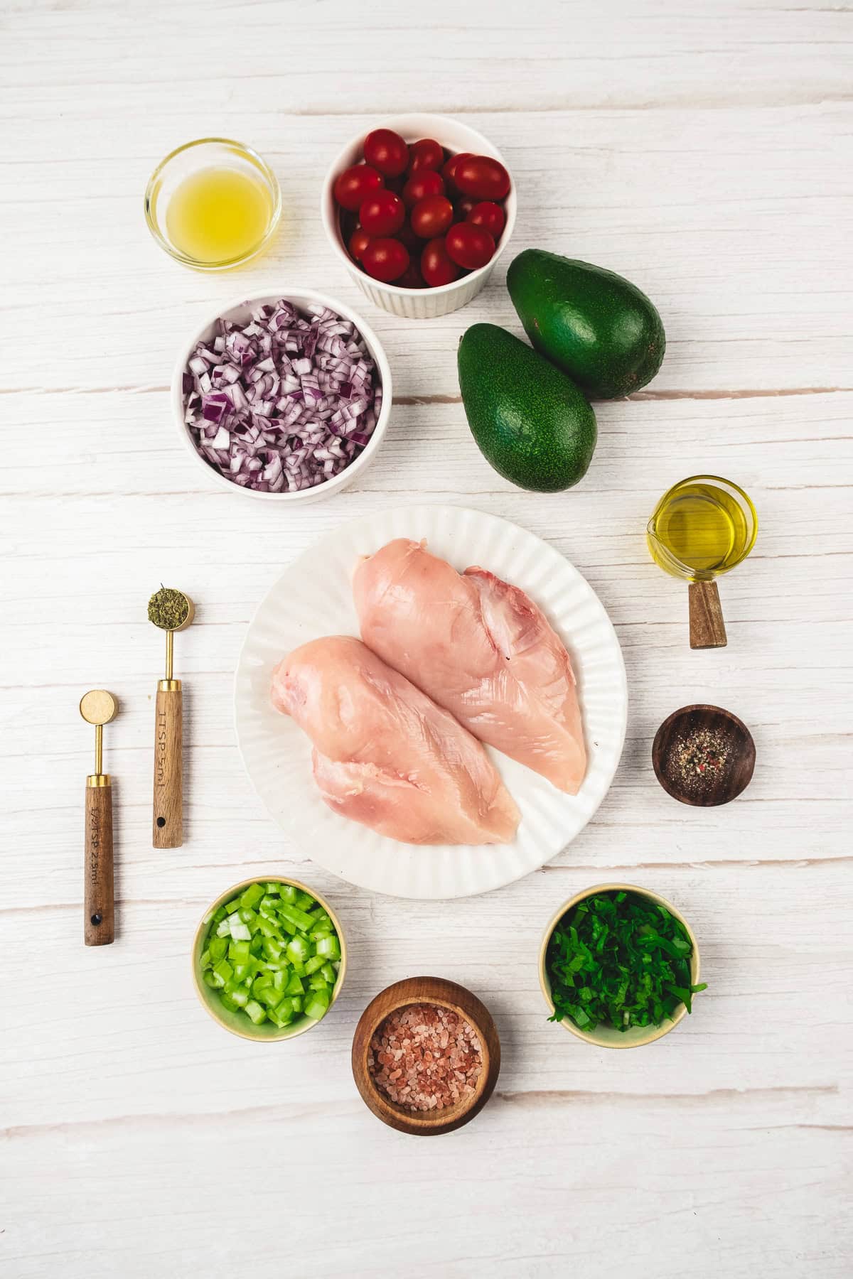 Two raw chicken breasts on a plate are surrounded by avocado, tomatoes, onion, celery, herbs, oil, and seasonings—perfect ingredients to craft a fresh Chicken Salad recipe.
