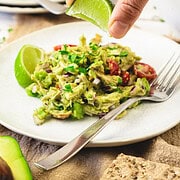 A hand squeezes a lime wedge over a plate of fresh avocado guacamole with tomatoes, onions, and herbs.